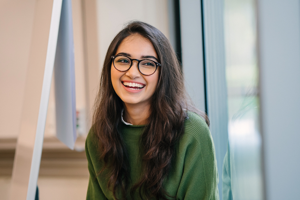 A close up head shot portrait of a preppy, young, beautiful, confident and attractive Indian Asian woman in a green sweater and spectacles in a classroom or office. She is smiling happily. 